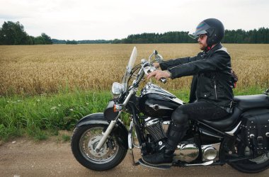 Bulutlu bir yaz gününde, arka planda kasklı, klasik siyah motosikletli bir adam.