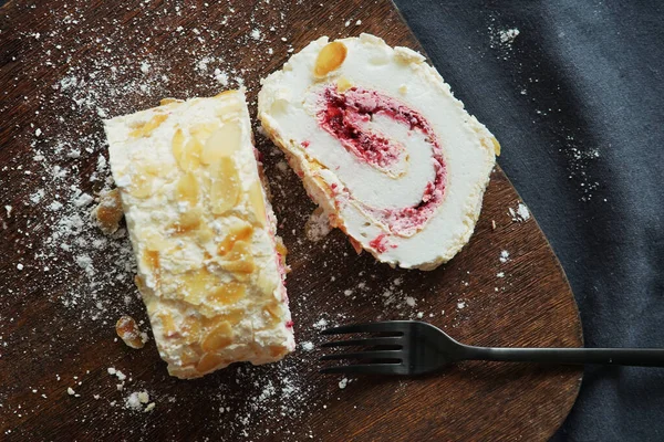 Meringue roll with raspberry jam and almonds next to a fork on a wooden board on a dark tablecloth