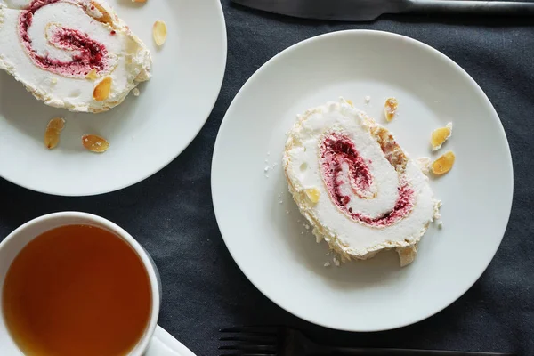 Pieces of meringue roll with raspberry jam and almonds on white plates next to a cup of tea on a gray tablecloth