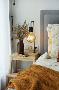 Pencerenin yanında lamba ve dekor olan ahşap masa ve kahverengi ekose bir yatak.