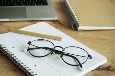 Açık bir dizüstü bilgisayar, defterler, bir kalem ve gözlük tahta bir masanın üzerinde duruyor.