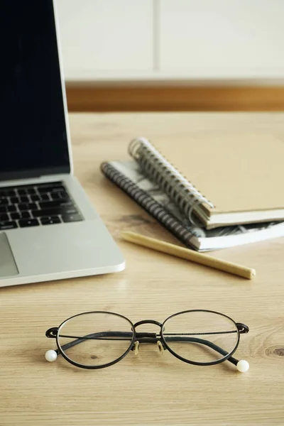 An open laptop, notebooks, a pen and glasses lie on a wooden table