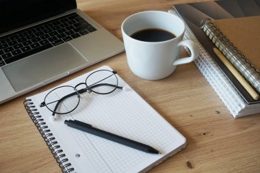 Kalem ve gözlüklü not defteri, dosyalar, dizüstü bilgisayar ve kahve fincanı ahşap masanın yanında.
