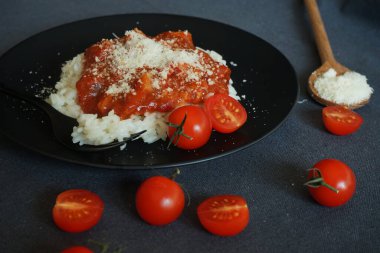 Domates soslu pilav, köfte ve siyah tabakta parmesan peyniri yanında çatal ve kiraz domatesli gri masa örtüsü.