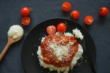 Domates soslu pilav, köfte ve siyah tabakta parmesan peyniri yanında çatal ve kiraz domatesli gri masa örtüsü.