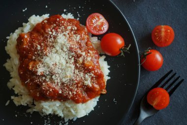 Domates soslu pilav, köfte ve siyah tabakta parmesan peyniri yanında çatal ve kiraz domatesli gri masa örtüsü.