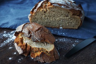 Taze ekmek parçalarının yanında bir somun ekmek ve tahtada siyah bir bıçak ve gri bir masa örtüsü var.