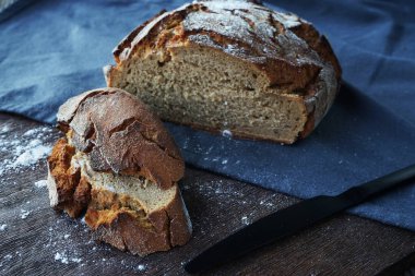 Taze ekmek parçalarının yanında bir somun ekmek ve tahtada siyah bir bıçak ve gri bir masa örtüsü var.