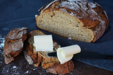 Taze ekmeğin üzerinde tereyağı parçaları var, ve yanında siyah bir bıçak, bir somun ekmek ve gri bir masa örtüsü var.