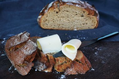 Taze ekmeğin üzerinde tereyağı parçaları var, ve yanında siyah bir bıçak, bir somun ekmek ve gri bir masa örtüsü var.