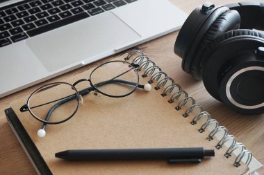 Siyah kulaklıklar, not defterleri, gözlükler ve ahşap bir masada yan yana dizüstü bilgisayar.