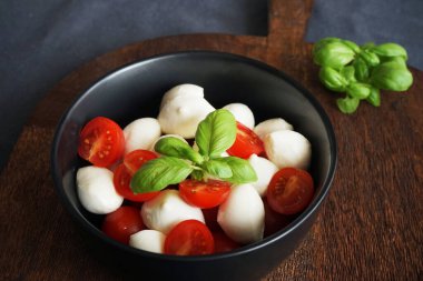 Siyah bir tabakta kiraz domatesli Mozzarella. Koyu gri arka planda ahşap bir tahtada fesleğen var.