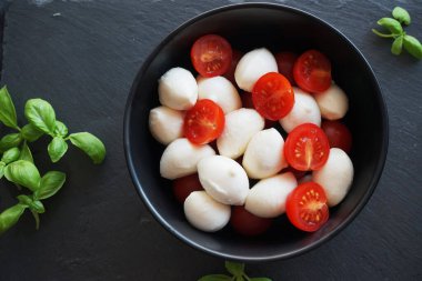 Koyu gri arka planda fesleğenli siyah bir tabakta kiraz domatesli Mozzarella.