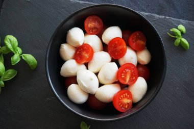 Koyu gri arka planda fesleğenli siyah bir tabakta kiraz domatesli Mozzarella.