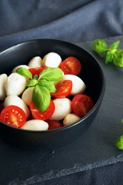 Koyu gri arka planda fesleğenli siyah bir tabakta kiraz domatesli Mozzarella.