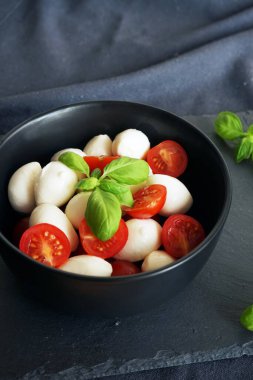 Koyu gri arka planda fesleğenli siyah bir tabakta kiraz domatesli Mozzarella.