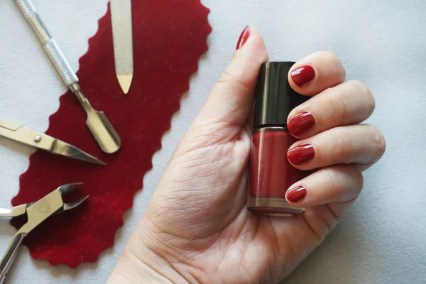 A woman\'s hand with painted nails holds a red polish next to manicure tools
