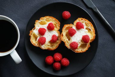 Kremalı ve ahududulu kekler siyah bir tabakta, bir fincan kahvenin yanında ve koyu arkaplanda bir bıçak.