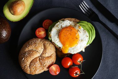 Yağda yumurta, avokado, siyah arka planda siyah bir tabakta ekmekli kiraz domatesleri.