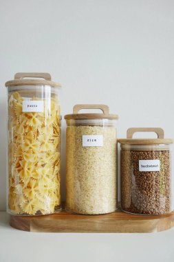 Beyaz zemin üzerinde makarna, pirinç ve karabuğday bulunan şeffaf kaplar.