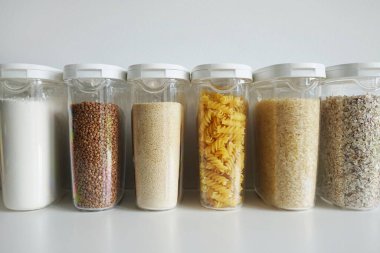 Beyaz zemin üzerinde makarna, pirinç, karabuğday, yulaf ezmesi ve un bulunan şeffaf kaplar.