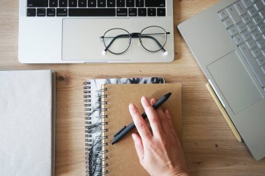 Bir el, bir defterin yanında bir kalem ve ahşap bir masanın üzerinde iki açık defter tutuyor.