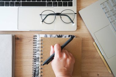 Bir el, bir defterin yanında bir kalem ve ahşap bir masanın üzerinde iki açık defter tutuyor.