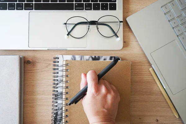 Bir el, bir defterin yanında bir kalem ve ahşap bir masanın üzerinde iki açık defter tutuyor.