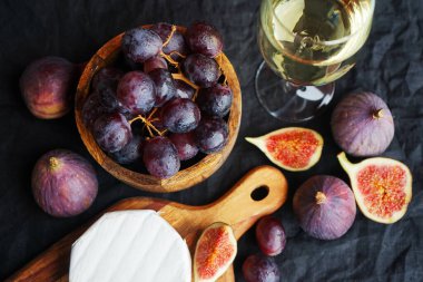 Camembert peyniri, olgun incir, kırmızı üzüm ve koyu arkaplanda bir bardak beyaz şarap.