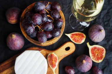 Camembert peyniri, olgun incir, kırmızı üzüm ve koyu arkaplanda bir bardak beyaz şarap.