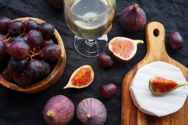 Camembert peyniri, olgun incir, kırmızı üzüm ve koyu arkaplanda bir bardak beyaz şarap.