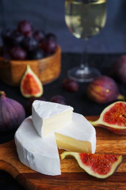 Camembert peyniri, olgun incir, kırmızı üzüm ve koyu arkaplanda bir bardak beyaz şarap.
