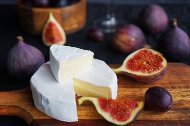 Camembert peyniri, olgun incir, kırmızı üzüm ve koyu arkaplanda bir bardak beyaz şarap.