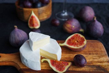 Camembert peyniri, olgun incir, kırmızı üzüm ve koyu arkaplanda bir bardak beyaz şarap.