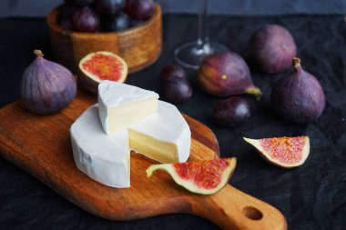 Camembert peyniri, olgun incir, kırmızı üzüm ve koyu arkaplanda bir bardak beyaz şarap.