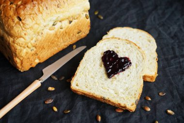 Koyu arkaplanda reçel kalpli bir parçanın yanında balkabağı tohumlu taze beyaz ekmek.