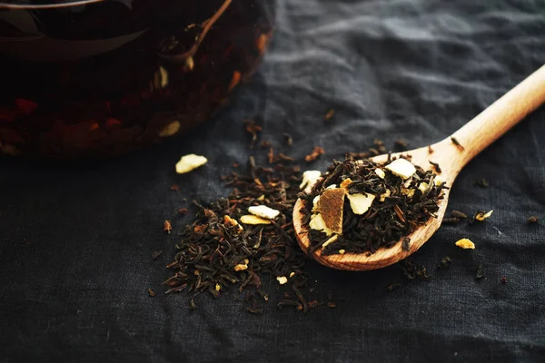 stock image Black tea with pieces of fruit on a wooden spoon next to the teapot on a dark background