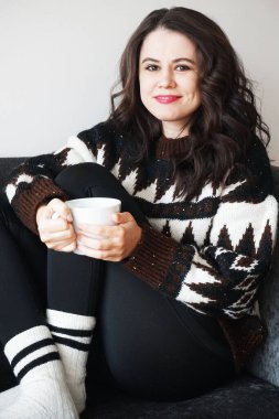 Koyu renk saçlı, etek desenli ve siyah taytlı bir kız elinde bir fincan çayla kanepede oturuyor.