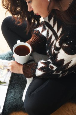 Koyu renk saçlı, etek desenli ve siyah taytlı, rahat kazaklı bir kız elinde bir fincan çayla bir kitabın yanında oturuyor.