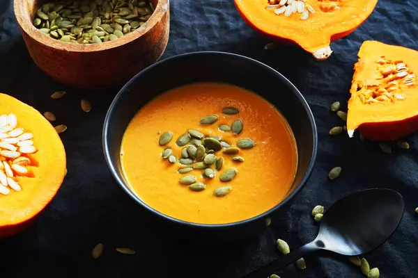 Pumpkin soup with pumpkin seeds next to a cut pumpkin on a dark background