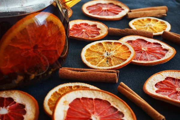 Glass teapot with black tea with orange next to dried orange and grapefruit slices on a dark background