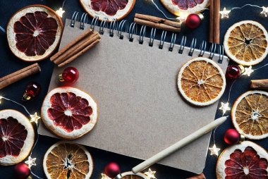 Kuru meyve dilimlerinin yanındaki kalemli not defteri, Noel ağacı süslemeleri, tarçın çubukları koyu arkaplanda.