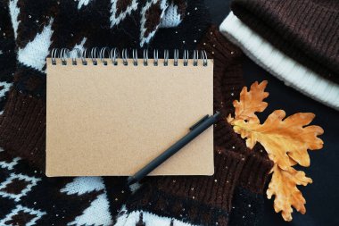 Siyah kalemli not defteri rahat bir süveterin üzerinde, şapka ve meşe yapraklarının yanında.