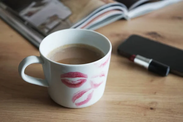 White cup of coffee with traces of lipstick on the background of a phone, book and red lipstick