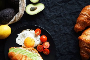 Avokado ile kruvasan, çırpılmış yumurta ve kiraz domatesleri siyah tabakta sebzelerin yanında ve arka planda bir çatal.