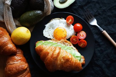 Avokado ile kruvasan, çırpılmış yumurta ve kiraz domatesleri siyah tabakta sebzelerin yanında ve arka planda bir çatal.