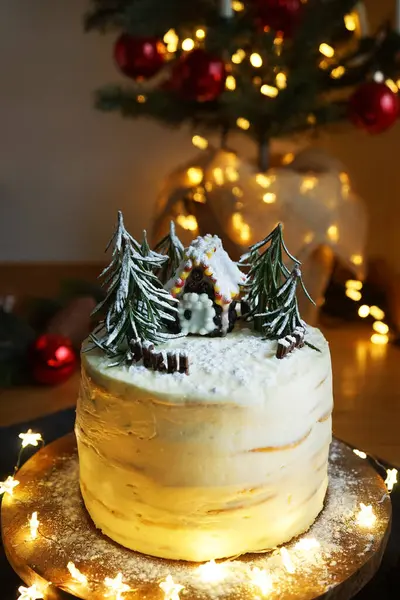 High cake with white cream with gingerbread house and fir trees on a background of Christmas lights