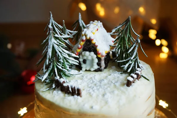 High cake with white cream with gingerbread house and fir trees on a background of Christmas lights