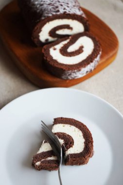 Tahtada vanilya kremalı çikolatalı dürüm, arka planda açık bir tabakta kesilmiş ruloların yanında.