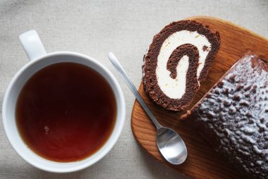 Çikolatalı sünger rulo vanilya kremalı tahta tahtada bir fincan çayın yanında.